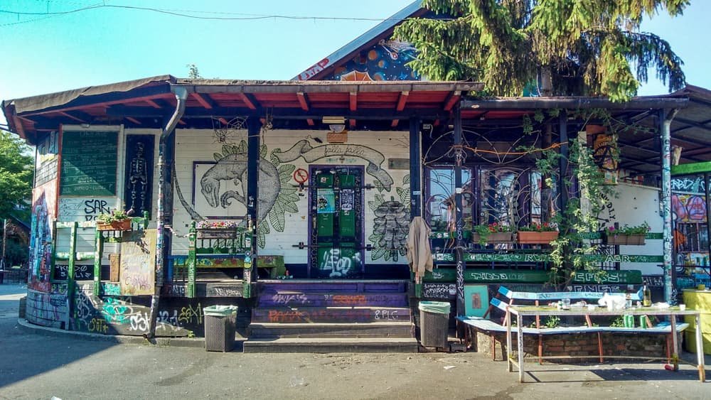 Uma casa com muitos grafites e pinturas em sua parede, além de plantas do lado lado de fora no bairro Metelkova, que fica na capital.