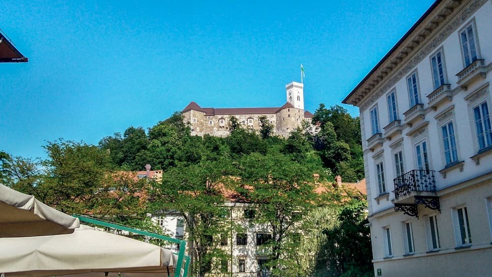 Castelo de Ljubljana cercado de vegetação visto no alto da colina pelo Mercado Central, em um dia de céu azul.