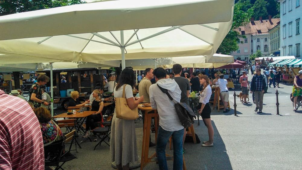 Mercado Central de Ljubljana com muitos turistas e locais espalhados por mesinhas e se deliciando com as comidas dos restaurantes nas tendinhas.