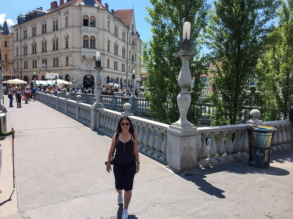 Marjorie atravessando a pé uma das Três Pontes (Ponte Tripla) na Praça Prešeren, em Ljubljana, em um dia de sol com muito movimento de turistas e locais atrás.