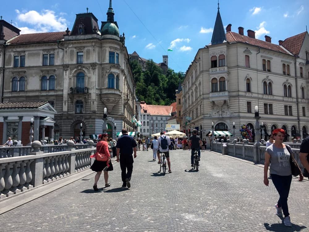 Pedestres andando pela Ponte Tripla com vista para o Castelo de Ljubljana ao fundo.