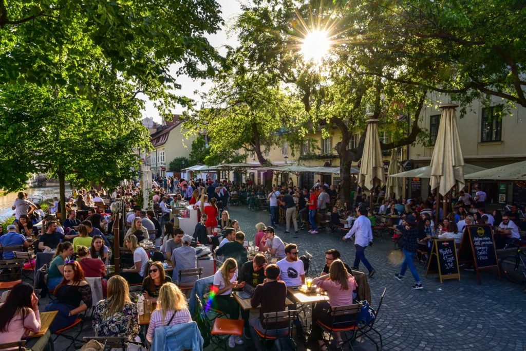 Rua Petkovškovo nabrežje, na cidade de Ljubljana, durante o dia e cheia de jovens ocupando as mesinhas (que ficam espalhadas na extremidade da rua) dos bares e restaurantes existentes por lá.