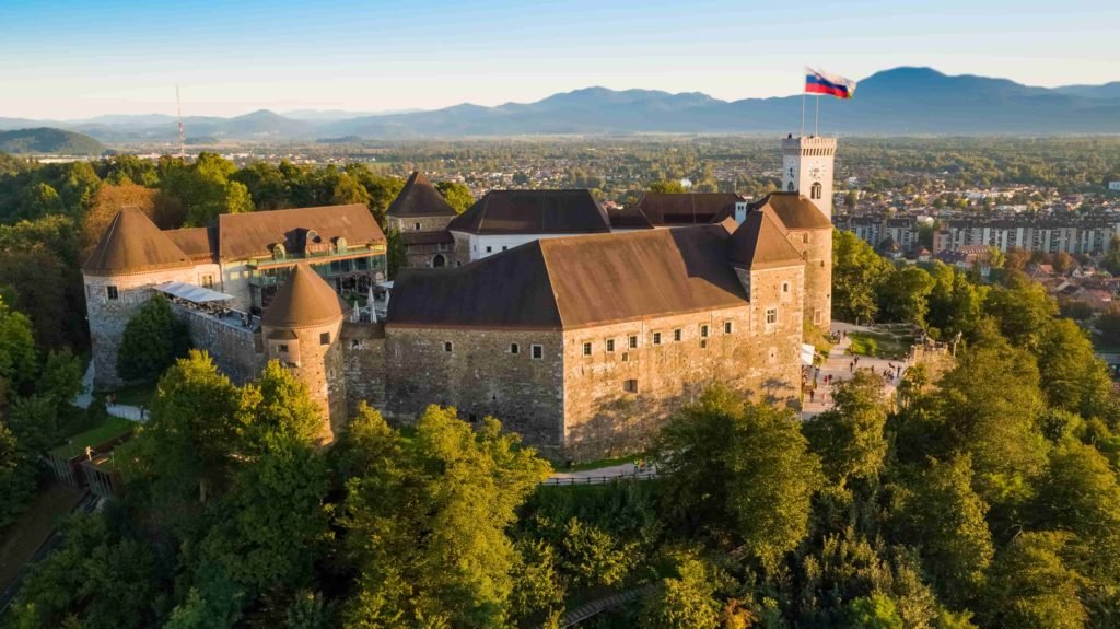 Vista aérea do Castelo de Ljubljana cercado de árvores e vegetação, em um dia ensolarado, com a bandeira balançando bem em cima de uma torre com relógio e o resto da cidade ao fundo.