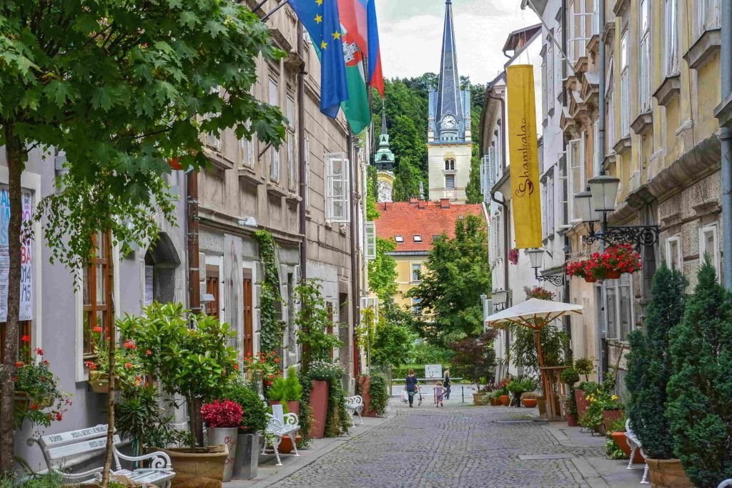 Uma das ruelinhas de pedras charmosas de Ljubljana, lotada de vasos de flores e plantas diferentes, com bandeiras penduradas nos prédios e uma torre de relógio ao fundo.