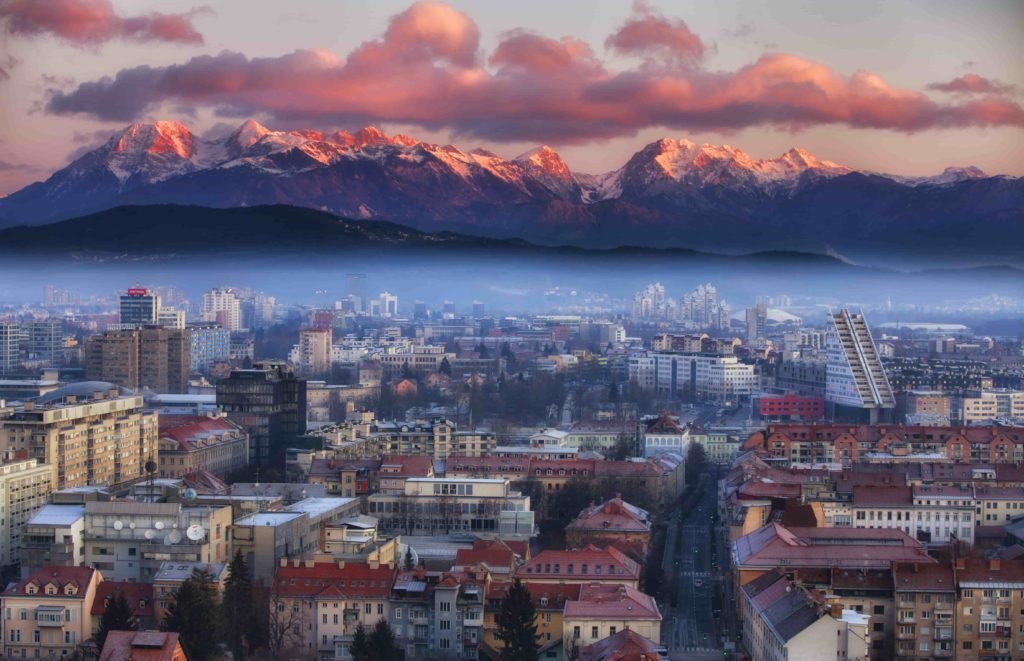 Vista de várias casas e prédios da cidade de Ljubljana, capital da Eslovênia, com montanhas ao fundo e muita neblina.