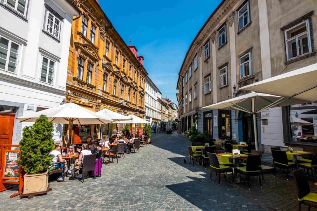 Stari Trg, também chamada de Old Square, uma avenida com muitos prédios que nos remetem à anos passados, com restaurantes e bares ocupando o térreo. Pessoas aproveitando o dia de sol sentadas em mesinhas, que ficam na rua de pedra, de um desses restaurantes.