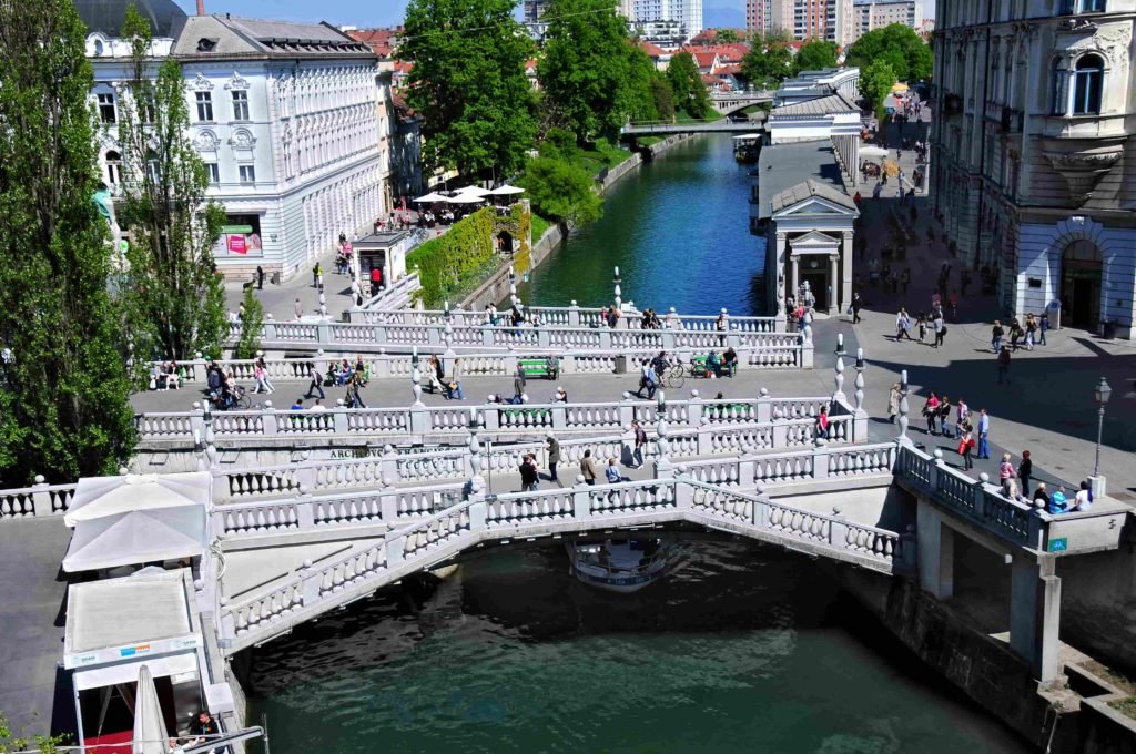 Vista aérea de pessoas transitando pela Ponte Tripla, que corta o Rio Ljubljanica, na Praça Prešeren, em um dia ensolarado e com barcos passando no Rio.