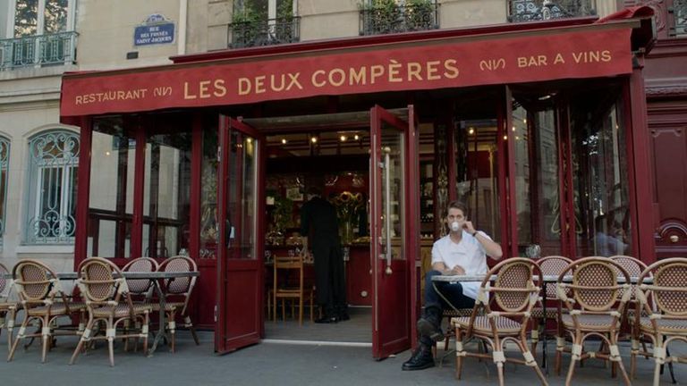Gabriel tomando um café sentado nas mesas que ficam lado de fora do restaurante de fachada vermelha que trabalha, o Les Deux Compères, que na vida real se chama Terra Nera.