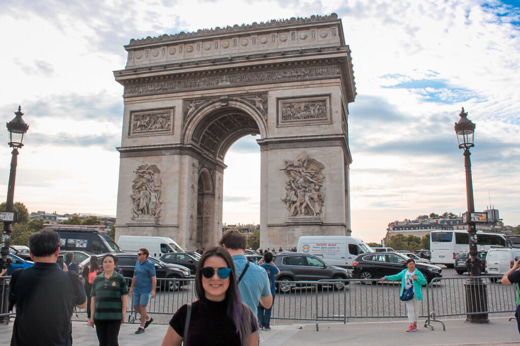 Marjorie e vários turistas tirando fotos com o Arco do Triunfo ao fundo, que fica bem no meio de uma rotatória. Na foto, só parte dessa rotatória aparece, e está com muitos carros passando, que nem quando Emily chega em Paris no primeiro episódio.