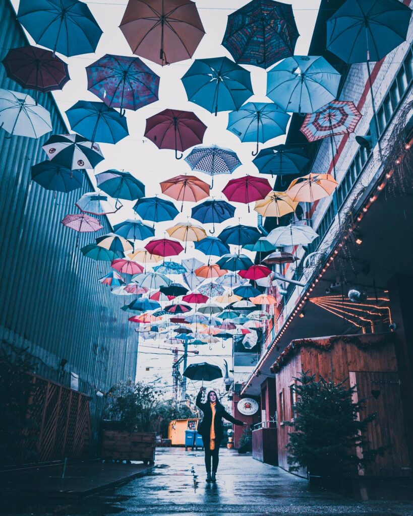 Marjorie segurando um guarda chuva debaixo de um teto de sombrinhas coloridas, que fica em um beco ao lado da Loja Freitag, no Zurich West.