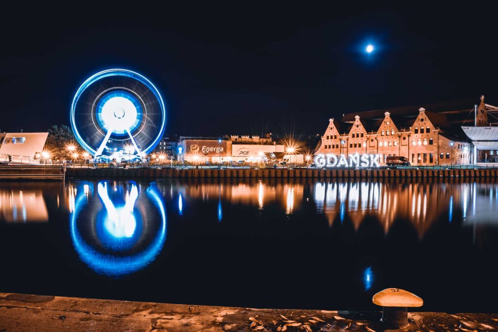 A Roda Gigante e o letreiro de Gdansk iluminados de azul e refletidos nas águas do Rio Motlawa.