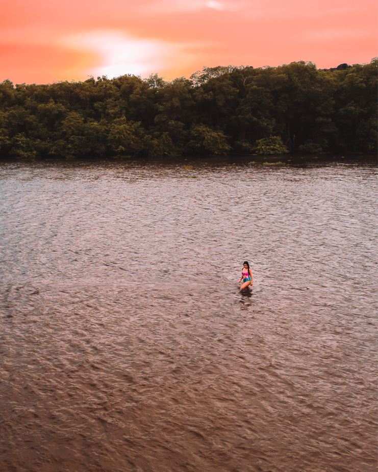 Marjorie andando na parte rasa do Rio Manguaba, com árvores o margeando ao fundo. O céu está rosa e laranja com o sol se pondo.